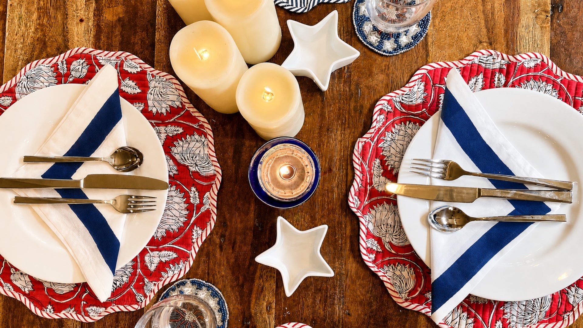 Dining table setting with red and white placemats, blue striped cloth napkins, candles, and cutlery on a wooden surface.