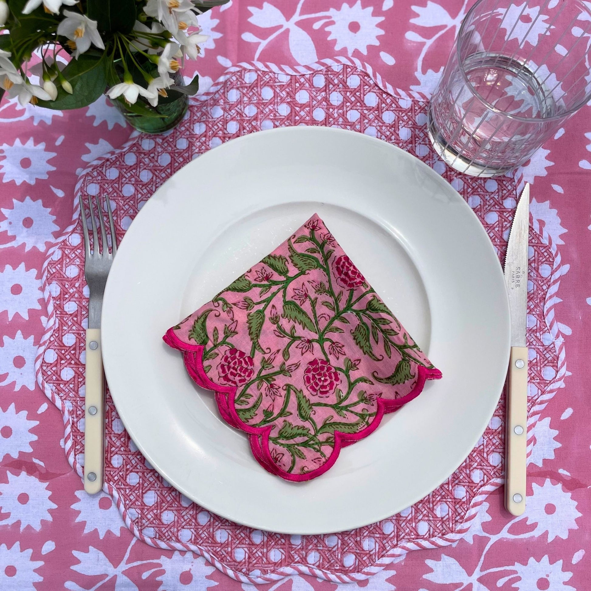 Table setting with a pink floral napkin on a white plate, placed on a pink and white patterned tablecloth.