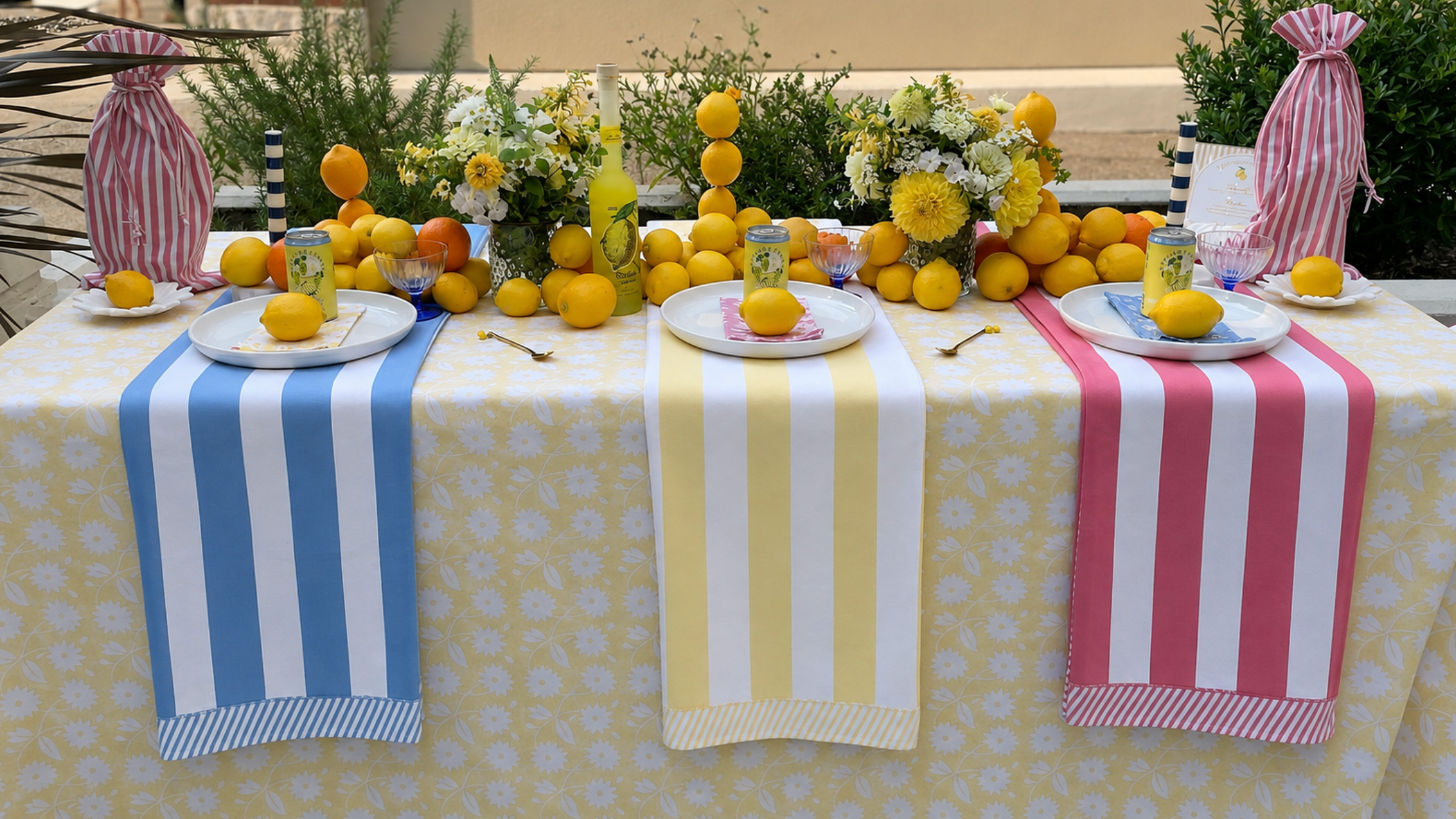Table set with yellow floral tablecloth and accented with blue stripe runner, yellow stripe runner and pink stripe runner, with real lemons and lemon drinks and yellow floral arrangements down the center of the table. 
